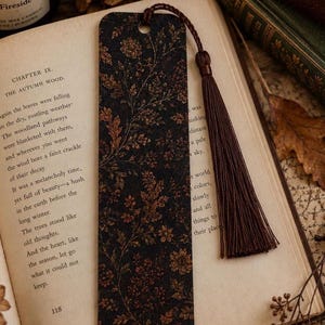 Moody autumn botanical bookmark with brown tassel resting on an open vintage book beside a glowing fall candle, antique books, dried flowers and autumn leaves in a cozy reading scene.