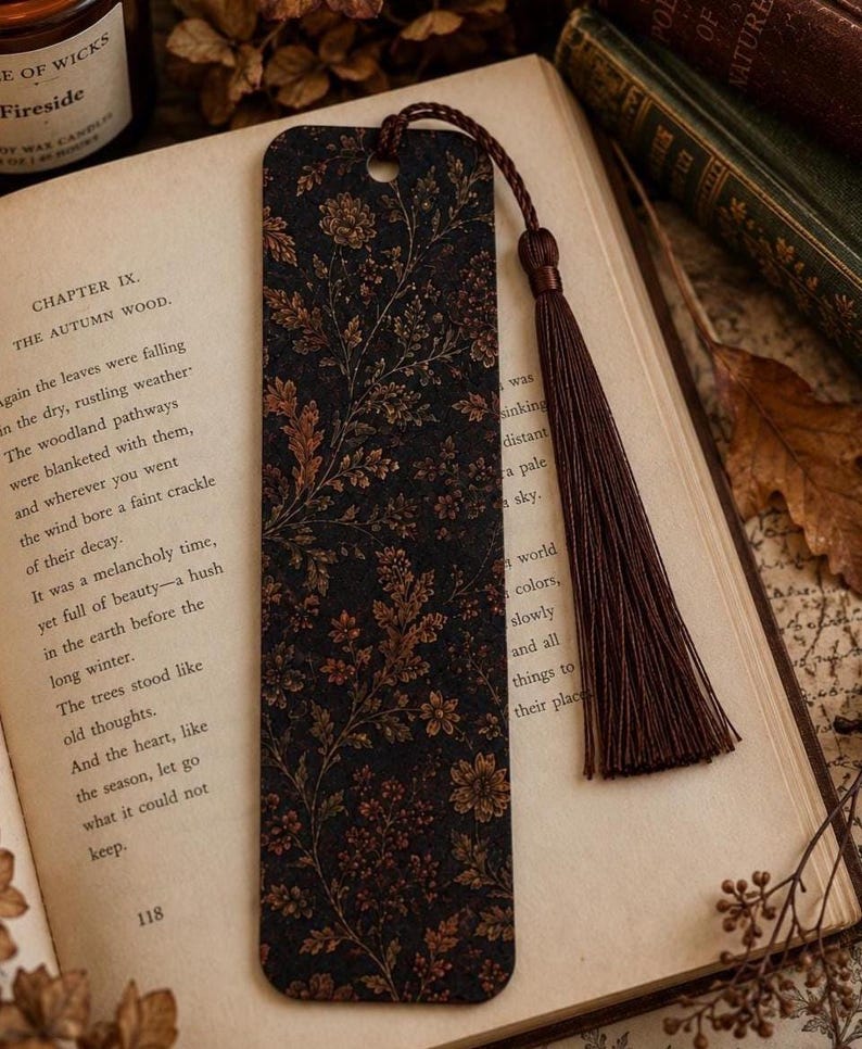 Moody autumn botanical bookmark with brown tassel resting on an open vintage book beside a glowing fall candle, antique books, dried flowers and autumn leaves in a cozy reading scene.