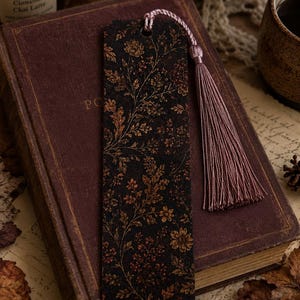 Moody autumn botanical bookmark with dusty mauve tassel displayed on a vintage burgundy book beside a lit fall candle, warm drink, dried flowers and autumn leaves in a cozy reading scene.