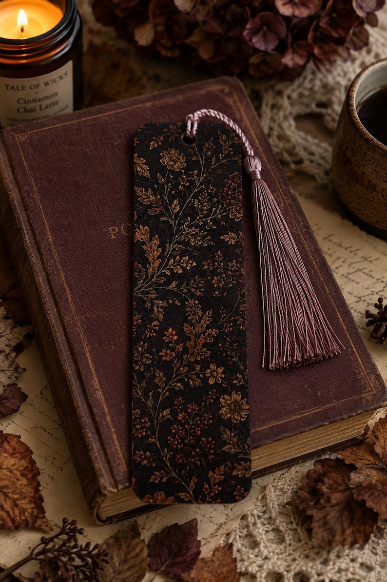 Moody autumn botanical bookmark with dusty mauve tassel displayed on a vintage burgundy book beside a lit fall candle, warm drink, dried flowers and autumn leaves in a cozy reading scene.