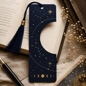 May include: A rectangular dark blue bookmark featuring a gold celestial print of stars, constellations, and moon phases. The bookmark has a large circular cutout on the right side and is finished with a dark blue tassel attached to the top.