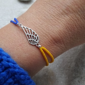 Könnte beinhalten: Ein Armband mit einem silbernen Flügel-Anhänger, aus blauem und gelbem Band.