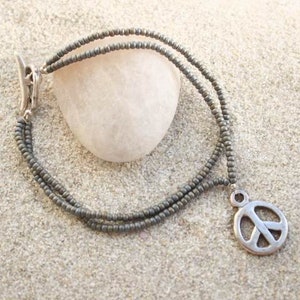 Könnte beinhalten: Eine graue Perlenkette mit einem silbernen Peace-Zeichen-Anhänger. Die Kette ist mehrlagig und hat einen silbernen Verschluss.