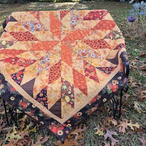 May include: A square quilt featuring a central star pattern composed of orange, yellow, and brown fabric patches. The design includes floral and leaf motifs, surrounded by a dark brown border decorated with pumpkins and sunflowers.