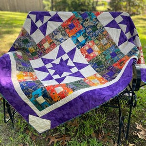 Peut inclure: Une courtepointe faite à la main drapée sur une chaise en métal noir. La courtepointe présente un motif en étoile avec des carrés violets, blancs et colorés. La bordure est en tissu violet uni.
