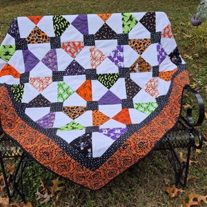 Może przedstawiać: Narzuta patchwork z powtarzającym się wzorem białych trójkątów i różnymi kwadratami tkanin o tematyce Halloween w kolorach pomarańczowym, fioletowym, czarnym i limonkowym. Narzuta wykończona jest szeroką pomarańczową lamówką w dynie.