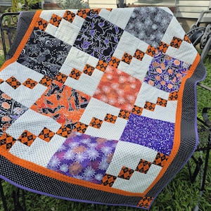 May include: A patchwork quilt featuring a Halloween theme with various fabric squares. The design includes patterns of witches, pumpkins, spiders, and webs in orange, purple, black, and white. The quilt is bordered by a black fabric with white dots.