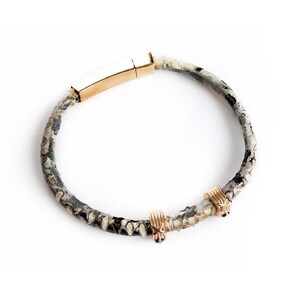 Könnte beinhalten: Ein Armband mit einem Schlangenlederband in Grau-, Schwarz- und Cremetönen. Es hat einen goldfarbenen rechteckigen Verschluss und zwei kleine goldfarbene dekorative Elemente. Das Armband ist zum Tragen am Handgelenk konzipiert.