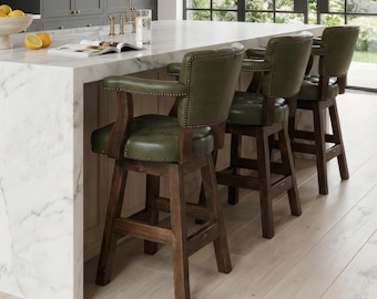 May include: A kitchen island with a white marble countertop and waterfall edge, featuring three dark wood bar stools with olive green tufted leather seats and brass nailhead trim, positioned under three cylindrical pendant lights.