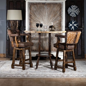 Op de afbeelding: Twee bruine houten barstoelen met een bruine leren zitting en een rugleuning met een gestoffeerd ontwerp. De krukken staan aan weerszijden van een ronde houten tafel met een wit marmeren blad. De tafel staat voor een witte kast met een bruin houtnerf ontwerp. De kast heeft twee deuren en is versierd met een witte rand. De kamer heeft een grijze muur en een tapijt met een lichtbruin en wit patroon.