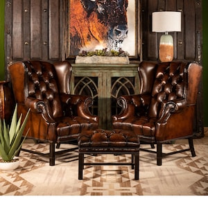 May include: Two brown leather wingback chairs with button tufting and a matching brown leather ottoman in front of a green cabinet with glass doors. A framed painting of a buffalo head is hanging on the wall behind the cabinet.