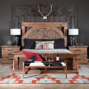 May include: A rustic wooden bed frame with a headboard featuring a large, arched panel. The bed is dressed with a black and white cowhide throw blanket and a red throw pillow. Two bedside tables with drawers are on either side of the bed. A wooden bench with a cowhide seat sits in front of the bed.