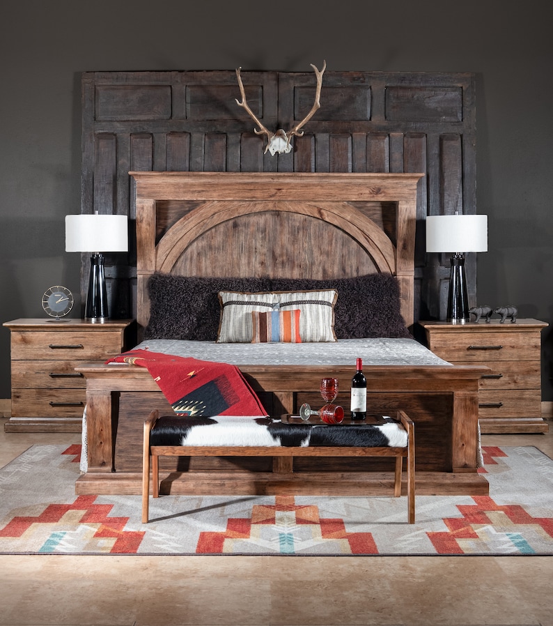 May include: A rustic wooden bed frame with a headboard featuring a large, arched panel. The bed is dressed with a black and white cowhide throw blanket and a red throw pillow. Two bedside tables with drawers are on either side of the bed. A wooden bench with a cowhide seat sits in front of the bed.