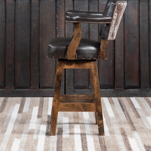 May include: A brown wooden bar stool with a dark brown leather seat and backrest. The backrest has a cowhide design. The stool has armrests and is adorned with small metal studs. The stool is set against a dark wooden door.