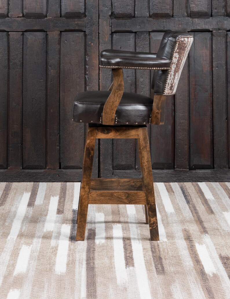 May include: A brown wooden bar stool with a dark brown leather seat and backrest. The backrest has a cowhide design. The stool has armrests and is adorned with small metal studs. The stool is set against a dark wooden door.