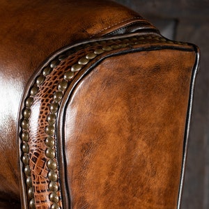 May include: Close-up of a brown leather armchair with a decorative stitched border and brass studs. The leather has a textured alligator pattern.