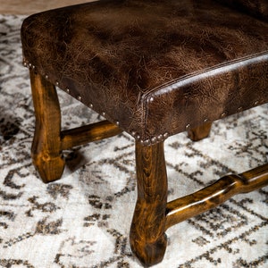 May include: Close-up of a brown leather chair with a dark wood frame. The seat is a distressed brown leather, and the frame is a dark, polished wood. The chair has decorative metal studs around the seat's perimeter. The chair is on a patterned rug.