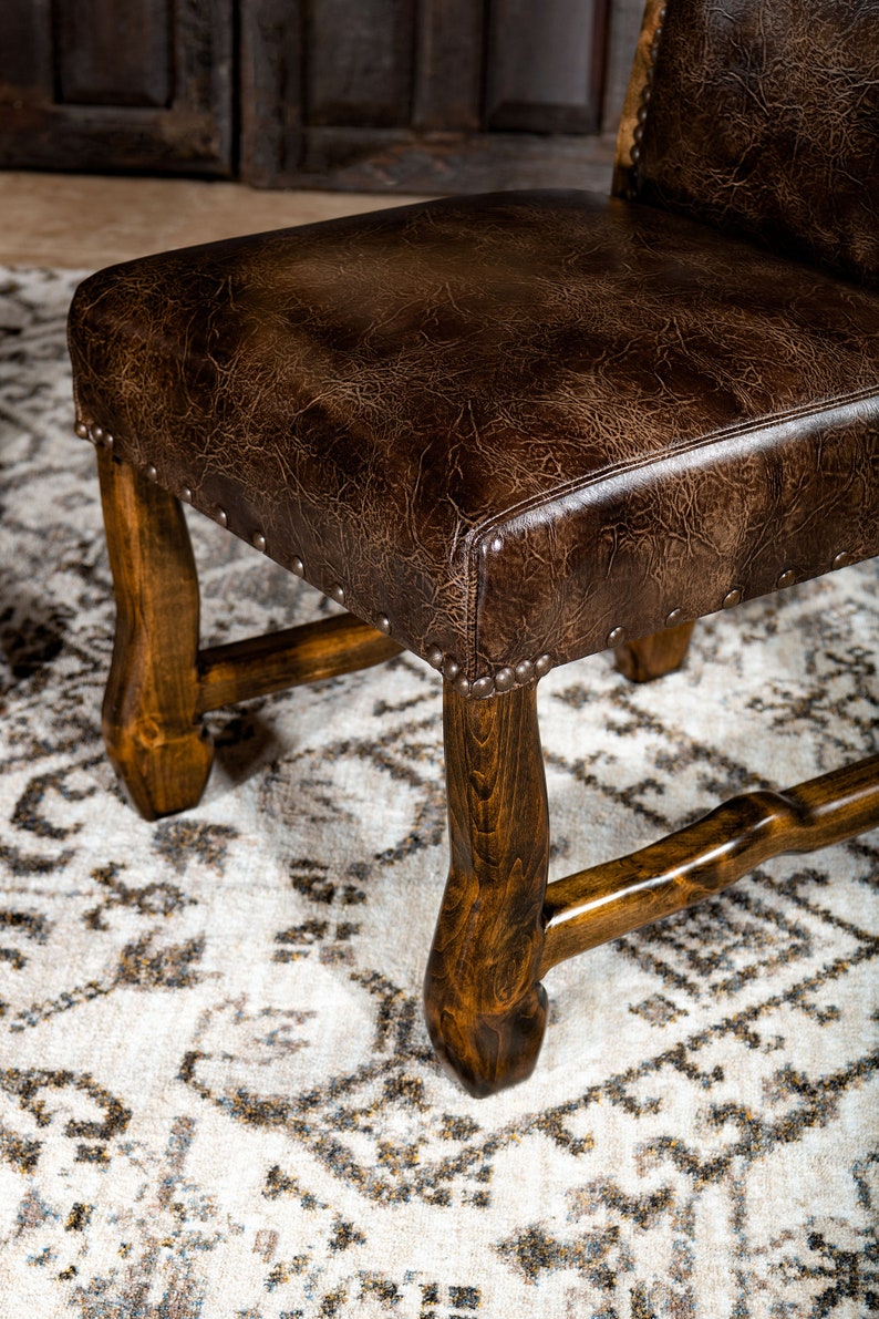 May include: Close-up of a brown leather chair with a dark wood frame. The seat is a distressed brown leather, and the frame is a dark, polished wood. The chair has decorative metal studs around the seat's perimeter. The chair is on a patterned rug.