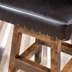 May include: Close-up of a brown wooden bar stool with a dark brown leather seat. The seat is square and has silver-colored nailhead trim. The wooden legs are a warm brown color.
