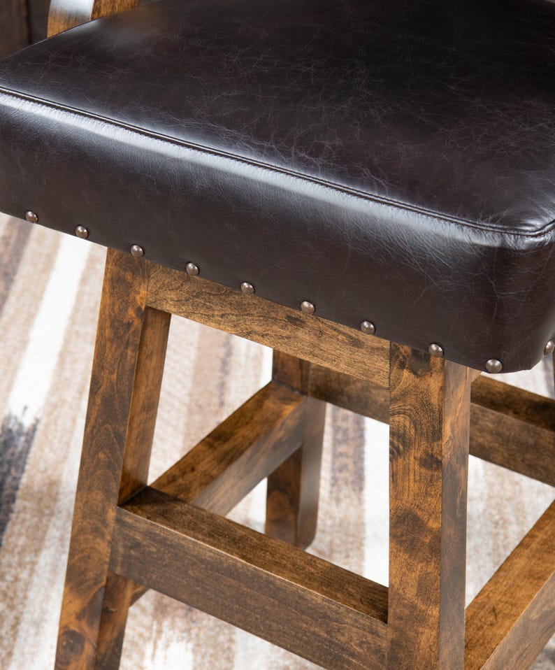 May include: Close-up of a brown wooden bar stool with a dark brown leather seat. The seat is square and has silver-colored nailhead trim. The wooden legs are a warm brown color.