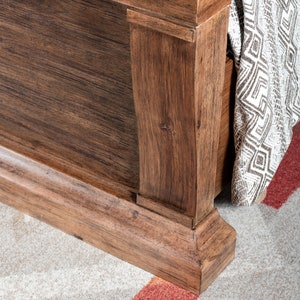 May include: Close-up of the bottom of a wooden bed frame with a decorative molding. The frame is a dark brown wood with a natural wood grain finish. The frame is sitting on a light beige carpet with a red and white patterned rug underneath.