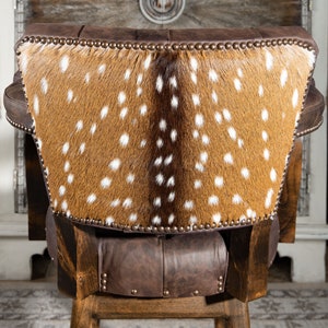 Op de afbeelding: Een bruine leren barkruk met een rugleuning bekleed met een gevlekt hertenhuid. De kruk heeft een houten frame en is versierd met metalen studs.