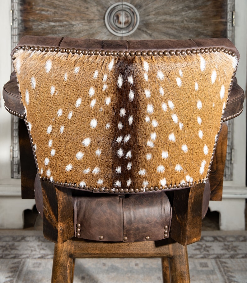 Op de afbeelding: Een bruine leren barkruk met een rugleuning bekleed met een gevlekt hertenhuid. De kruk heeft een houten frame en is versierd met metalen studs.