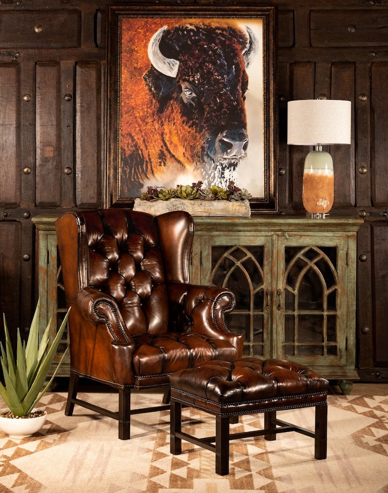 May include: A brown leather armchair with a tufted back and seat, and a matching ottoman. The chair and ottoman are in front of a green cabinet with glass doors. A painting of a buffalo is hanging on the wall behind the cabinet.