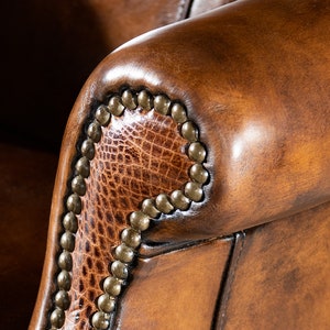 May include: Close-up of a brown leather armchair with a decorative nailhead trim. The armrest is made of a textured leather with a subtle pattern.
