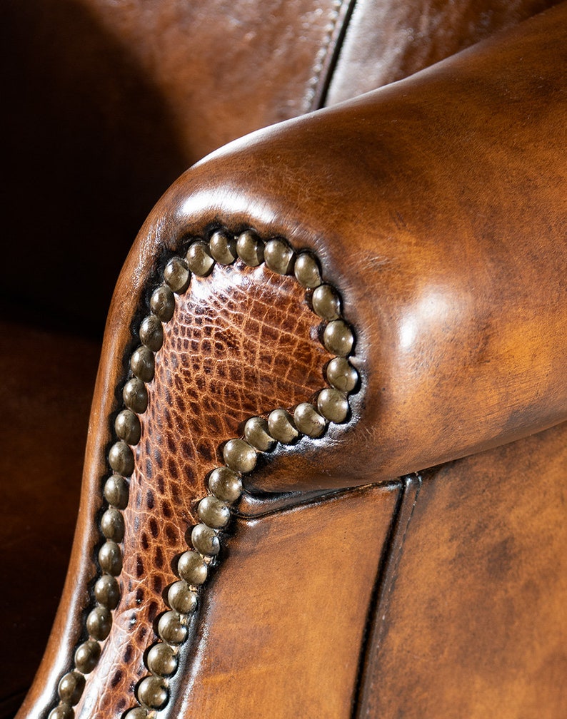 May include: Close-up of a brown leather armchair with a decorative nailhead trim. The armrest is made of a textured leather with a subtle pattern.
