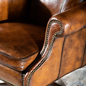 May include: Close-up of a brown leather office chair with a detailed, studded trim. The chair has a high back and a comfortable seat.
