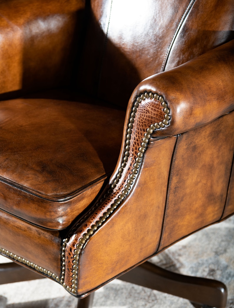 May include: Close-up of a brown leather office chair with a detailed, studded trim. The chair has a high back and a comfortable seat.