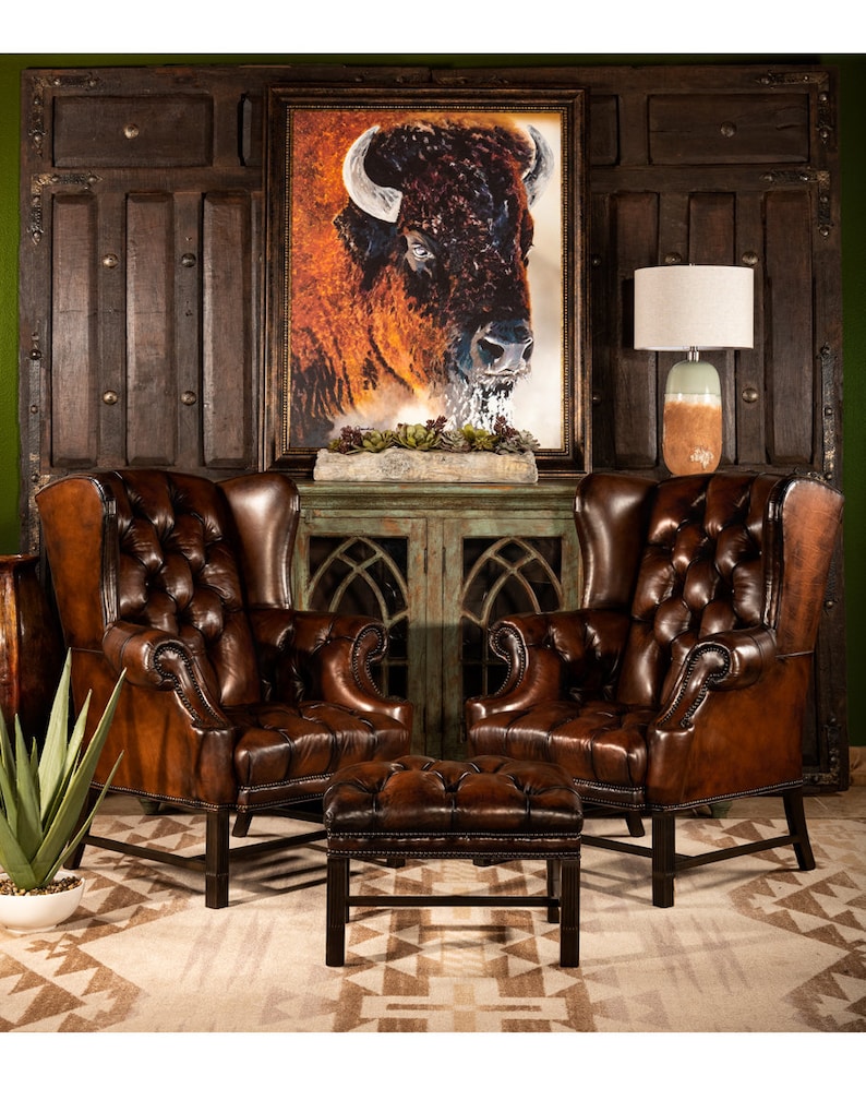 May include: Two brown leather wingback chairs with button tufting and a matching brown leather ottoman in front of a green cabinet with glass doors. A framed painting of a buffalo head is hanging on the wall behind the cabinet.