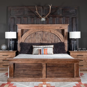 May include: A rustic wooden bed frame with a headboard featuring an arched design and a dark wood finish. The bed is dressed with a black faux fur throw and a colorful patterned pillow. Two bedside tables with drawers are visible on either side of the bed.