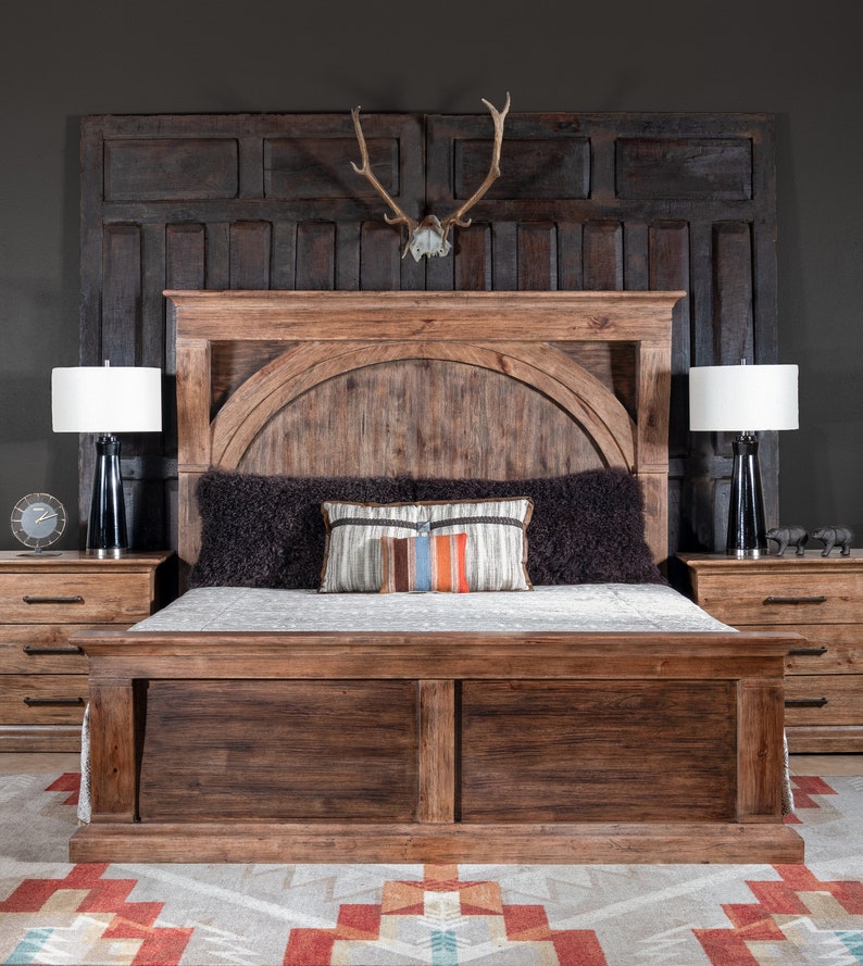 May include: A rustic wooden bed frame with a headboard featuring an arched design and a dark wood finish. The bed is dressed with a black faux fur throw and a colorful patterned pillow. Two bedside tables with drawers are visible on either side of the bed.