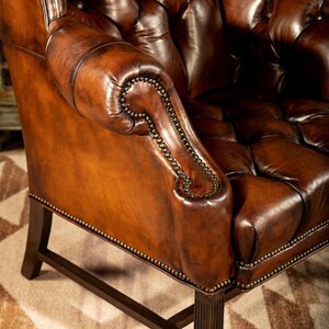 May include: A brown leather armchair with a tufted back and seat. The chair has a high back and rolled arms. The chair is upholstered in a rich brown leather and has a distressed finish. The chair is sitting on a rug with a geometric pattern.