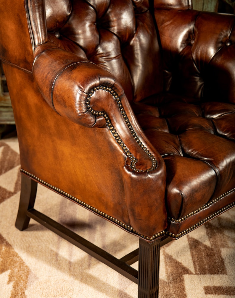 May include: A brown leather armchair with a tufted back and seat. The chair has a high back and rolled arms. The chair is upholstered in a rich brown leather and has a distressed finish. The chair is sitting on a rug with a geometric pattern.