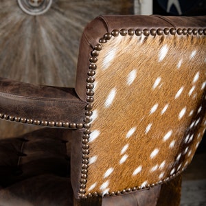 Op de afbeelding: Een bruine leren stoel met een rugleuning bekleed met een gevlekt koehuidpatroon. De stoel heeft een decoratieve spijkerkoprand rond de randen.