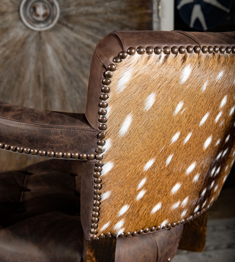 Op de afbeelding: Een bruine leren stoel met een rugleuning bekleed met een gevlekt koehuidpatroon. De stoel heeft een decoratieve spijkerkoprand rond de randen.