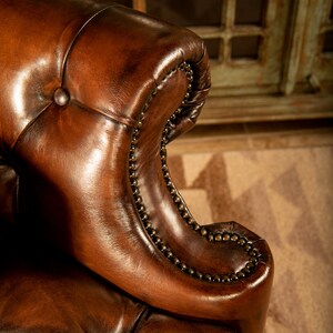 May include: Close-up of a brown leather armchair with button tufting and nailhead trim. The chair has a rolled back and a high, curved armrest.