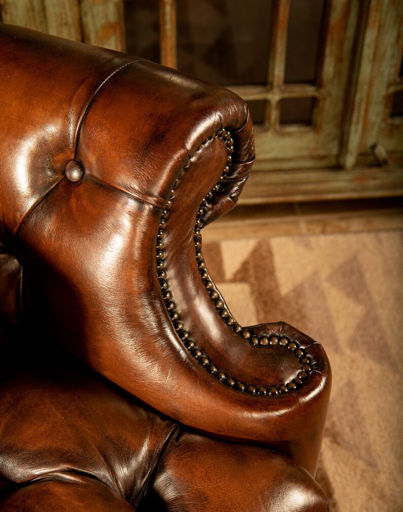 May include: Close-up of a brown leather armchair with button tufting and nailhead trim. The chair has a rolled back and a high, curved armrest.