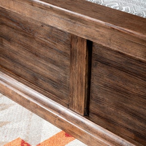 May include: Close-up of a wooden bed frame with a dark brown finish. The frame has a simple design with a raised edge and a vertical panel in the center.