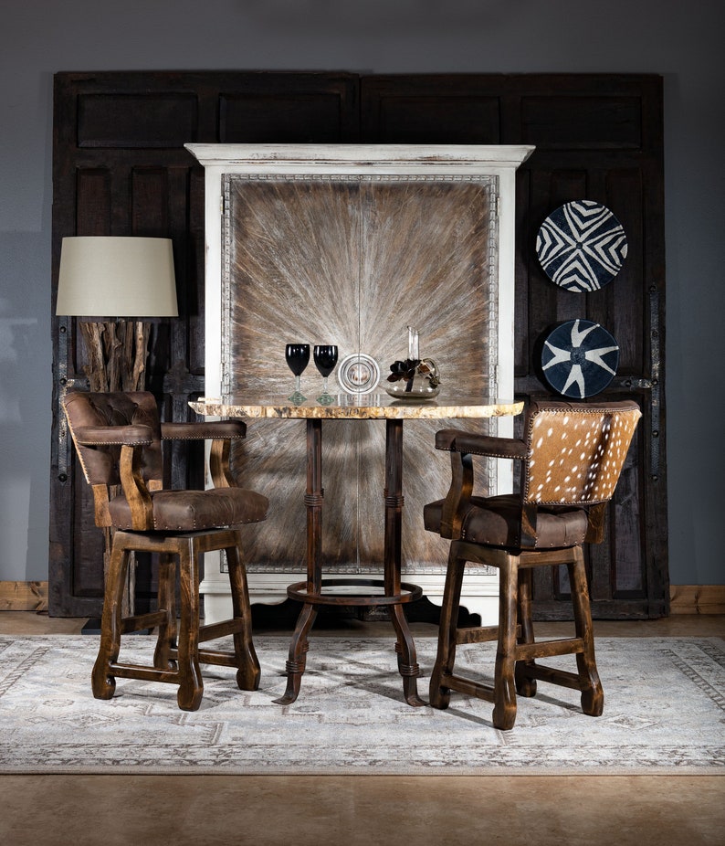 Op de afbeelding: Twee bruine houten barstoelen met een bruine leren zitting en een rugleuning met een gestoffeerd ontwerp. De krukken staan aan weerszijden van een ronde houten tafel met een wit marmeren blad. De tafel staat voor een witte kast met een bruin houtnerf ontwerp. De kast heeft twee deuren en is versierd met een witte rand. De kamer heeft een grijze muur en een tapijt met een lichtbruin en wit patroon.