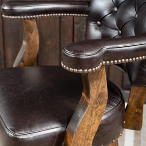 May include: Close-up of a brown leather armchair with button tufting on the backrest. The armrests are also brown leather, trimmed with small silver studs. The chair has wooden arm supports and a wooden base, creating a classic, vintage aesthetic.