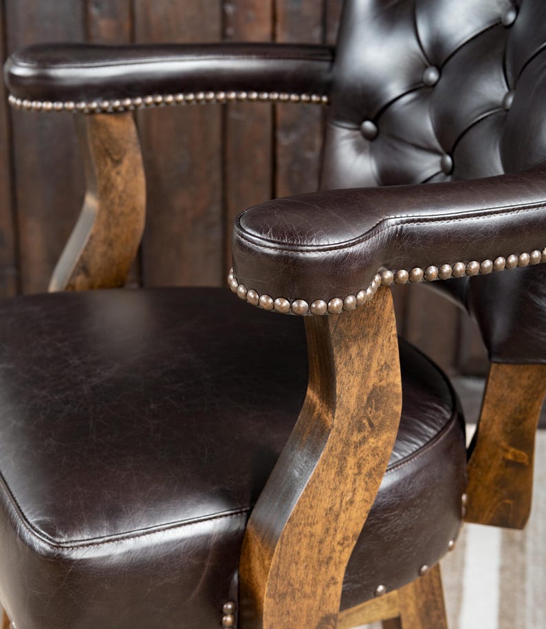 May include: Close-up of a brown leather armchair with button tufting on the backrest. The armrests are also brown leather, trimmed with small silver studs. The chair has wooden arm supports and a wooden base, creating a classic, vintage aesthetic.
