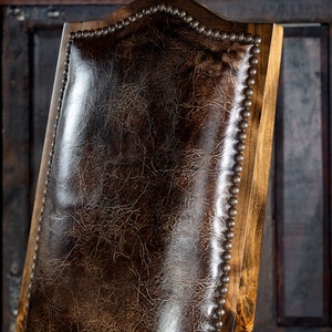 May include: Close-up of a wooden chair back with a dark brown leather seat. The leather has a textured, crackled appearance. The wooden frame is light brown, and the edges are adorned with small, evenly spaced metal studs.