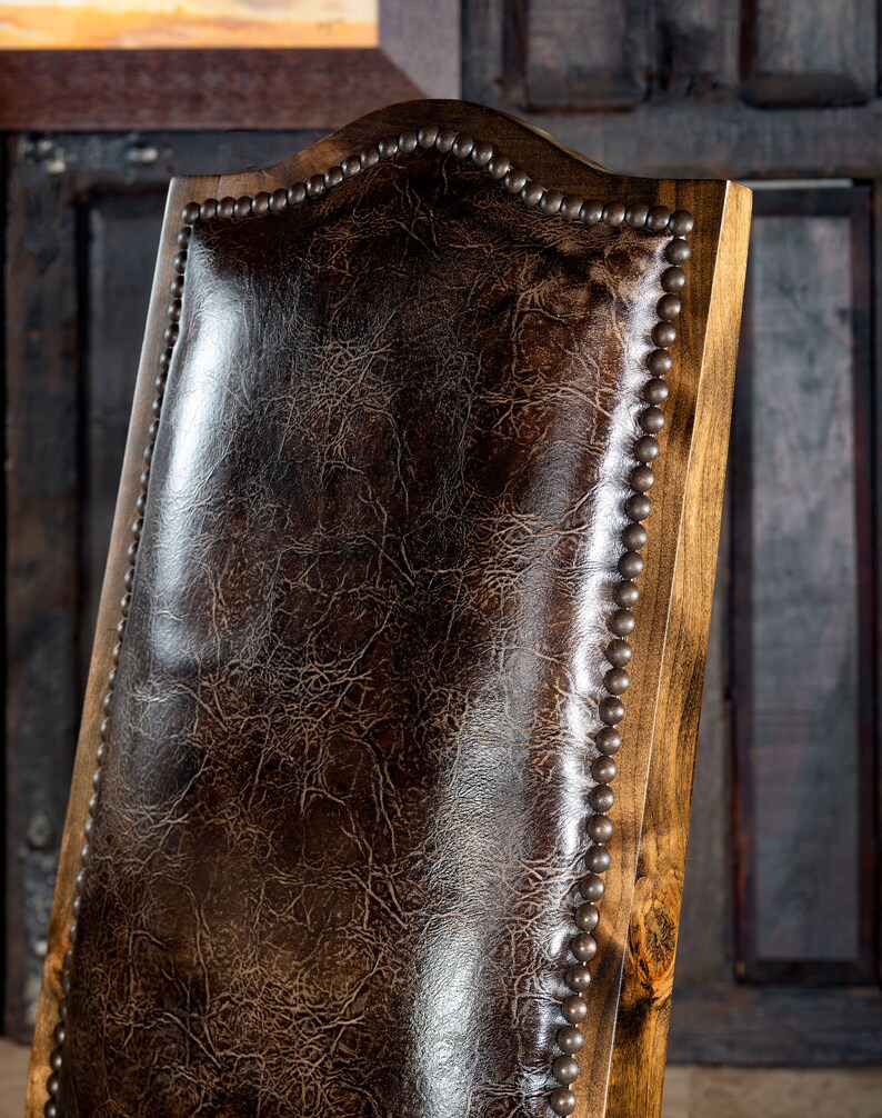 May include: Close-up of a wooden chair back with a dark brown leather seat. The leather has a textured, crackled appearance. The wooden frame is light brown, and the edges are adorned with small, evenly spaced metal studs.