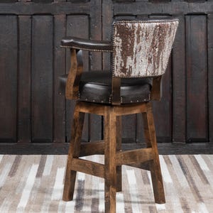 May include: A rustic bar stool with a dark brown leather seat and backrest. The backrest features a unique cowhide pattern in shades of brown and white. The wooden frame is a warm, medium brown, and the stool has armrests.
