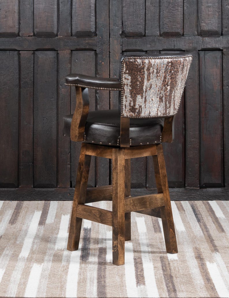May include: A rustic bar stool with a dark brown leather seat and backrest. The backrest features a unique cowhide pattern in shades of brown and white. The wooden frame is a warm, medium brown, and the stool has armrests.