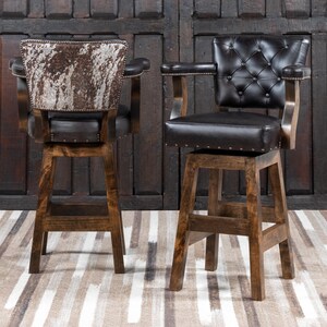 May include: Two wooden bar stools with dark brown leather seats and backs. One stool has a cowhide back, the other has a button-tufted back. Both have armrests and are on a light brown and white striped rug.